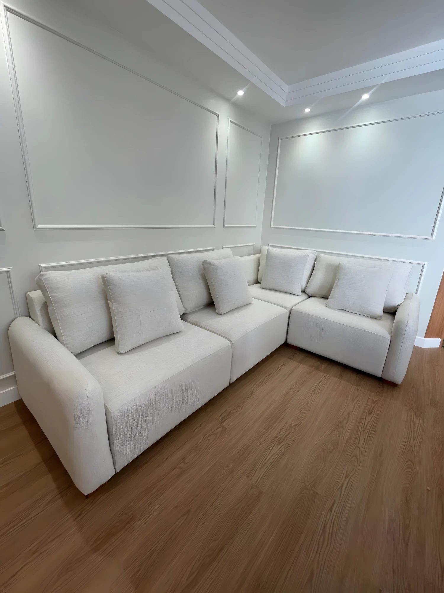 Sofá de canto off-white com almofadas, em sala com boiserie branca e piso de madeira